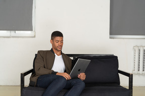 Front view of a bored young man sitting on a sofa while watching the tablet