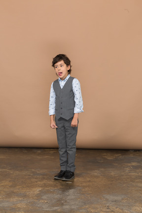 Vista frontal de un chico lindo en traje gris haciendo muecas y mostrando la lengua