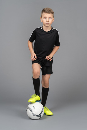 Front view of a child boy in football uniform putting his foot on ball and looking at camera