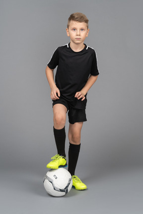 Vista frontal de un niño chico en uniforme de fútbol poniendo su pie en la pelota y mirando a la cámara