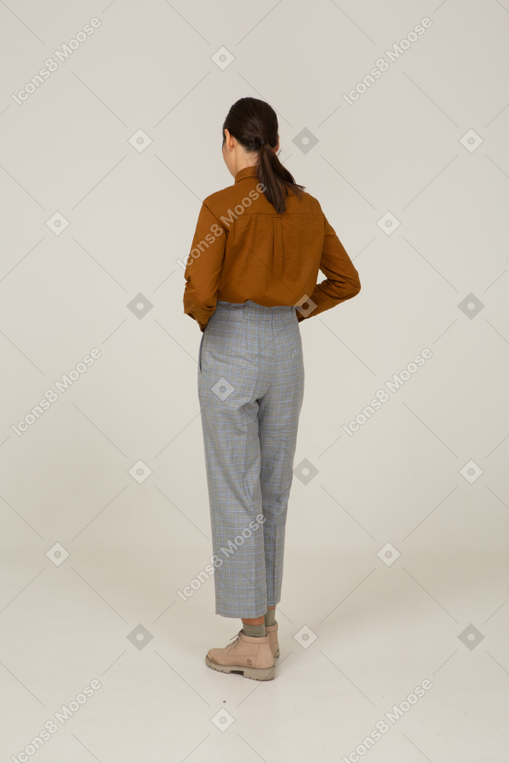Vista traseira a três quartos de uma jovem mulher asiática de calça e blusa tocando a barriga