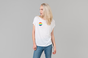 Gut aussehender junger mann mit langen blonden haaren, stehend vor grauem hintergrund, trägt blue jeans und ein weißes t-shirt mit lgbt-abzeichen darauf