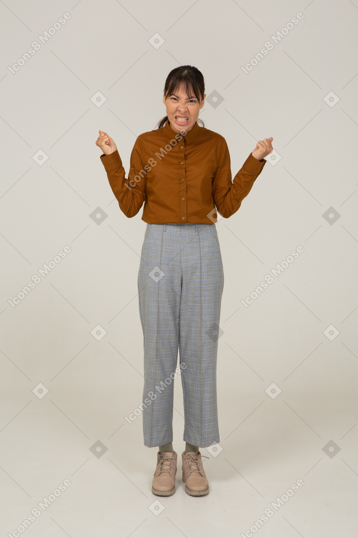Vista frontale di una giovane donna asiatica malvagia in calzoni e camicetta che stringe i denti e i pugni