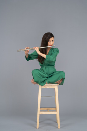 Toute la longueur d'une jeune femme jouant de la clarinette assise, les jambes croisées sur une chaise en bois