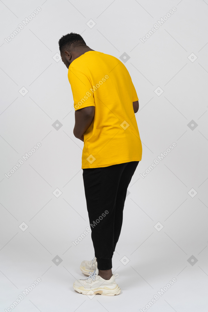 배를 만지고 아래를 내려다보는 노란색 티셔츠를 입은 검은 피부의 젊은 남자의 3/4 뒷모습