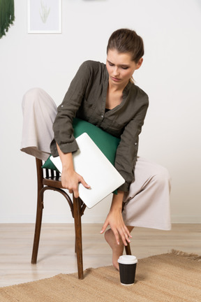Vista frontal de una mujer joven confundida sentada en una silla y sosteniendo su computadora portátil y tocando la taza de café