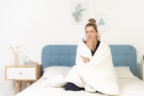 Front view of a yawning young woman in pajamas wrapped in blanket staying in bed