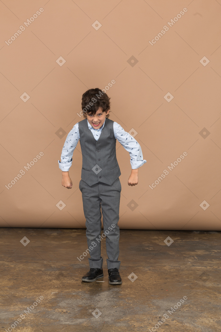 握りこぶしで立ってカメラを見ている灰色のスーツを着た怒っている少年の正面図