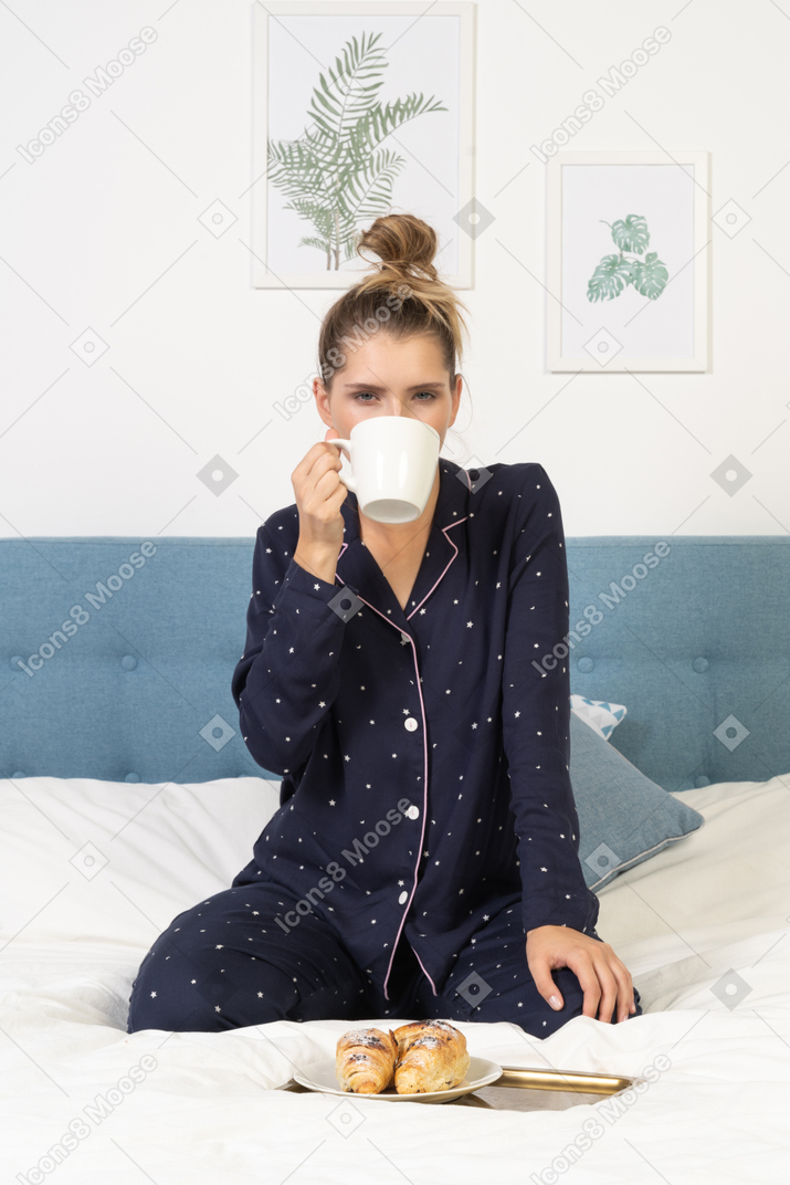 Front view of a young lady in pajamas drinking coffee in bed