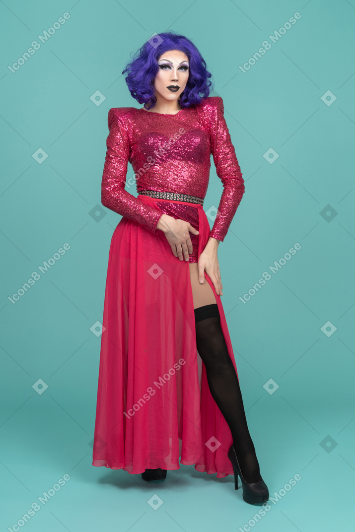 Vista frontal de uma drag queen de vestido rosa colocando um pé para frente e tocando a coxa