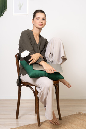 Вид в три четверти молодой женщины, сидящей на стуле, держащей ноутбук и трогающей чашку кофе