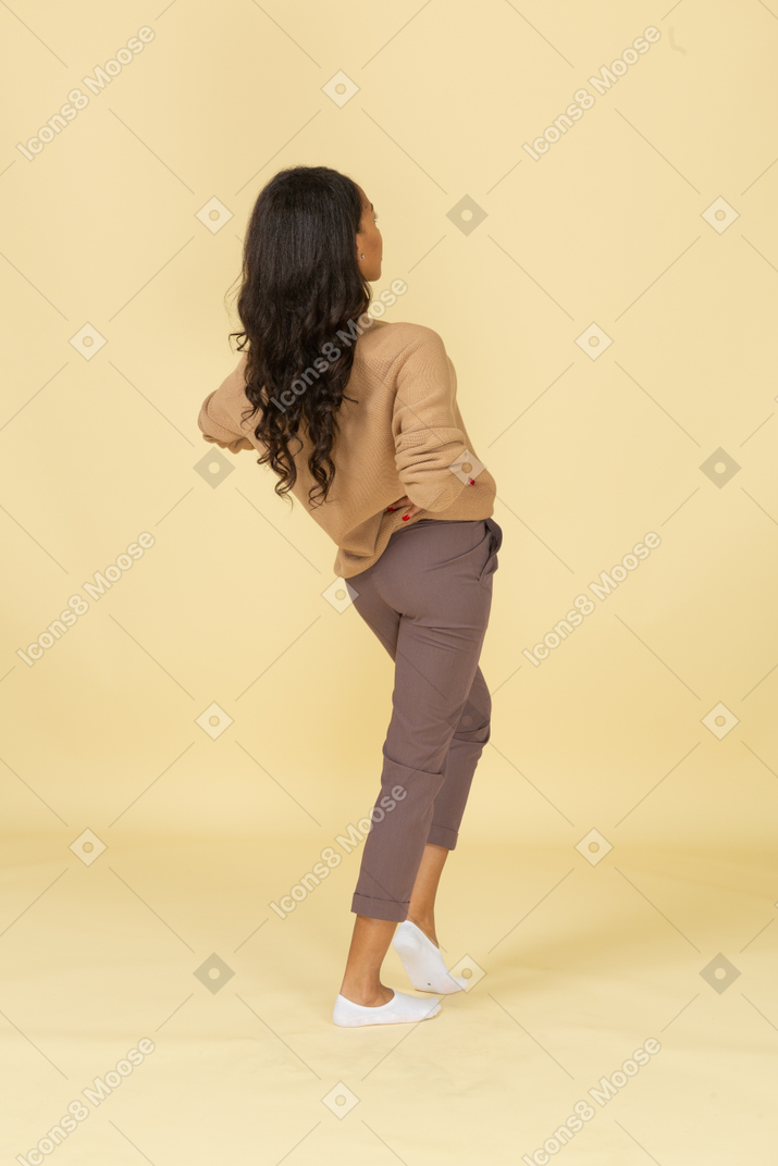 Vista posterior de tres cuartos de una mujer joven de piel oscura que pone las manos en las caderas y se inclina hacia atrás