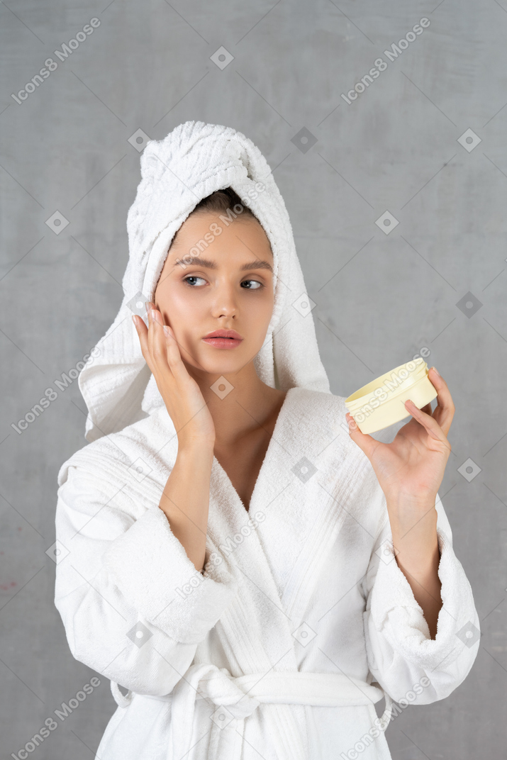 Woman in bathrobe touching cheek and holding a tub of cream