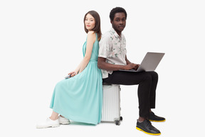 Interracial couple sitting on suitcase and and using gadjets