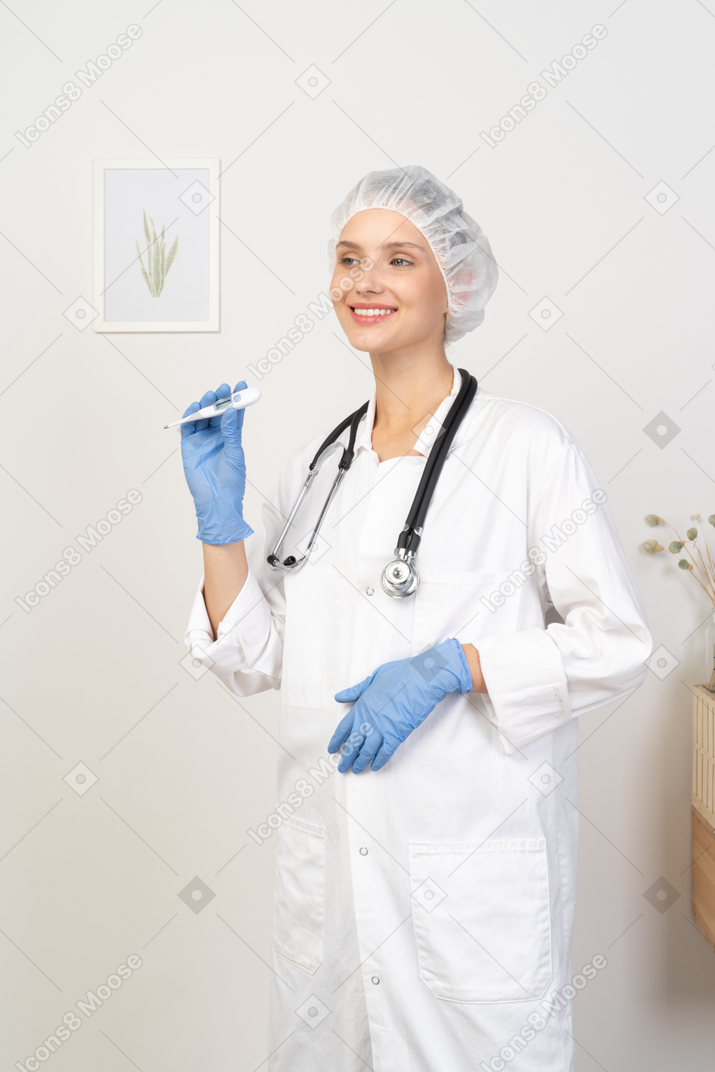 Vue de trois quarts d'une jeune femme médecin souriante avec stéthoscope tenant un thermomètre