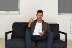 Vorderansicht eines jungen mannes, der auf einem sofa sitzt und eine zigarette hält und kaffee trinkt