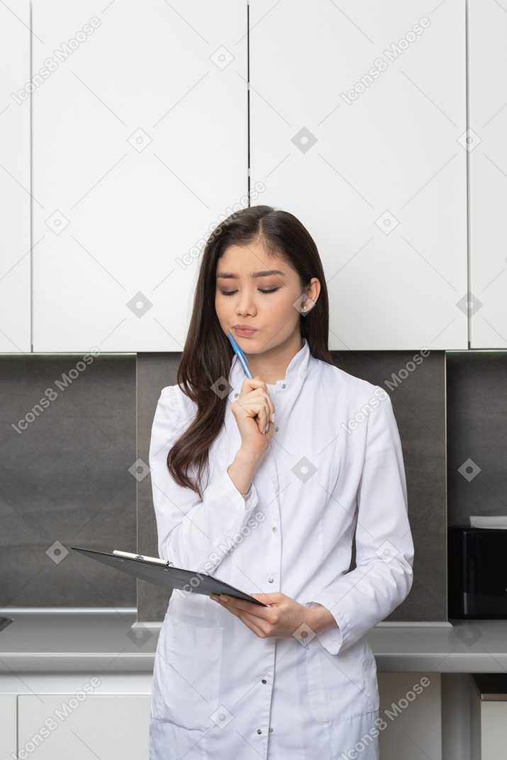 Front view of a female doctor touching face with a pen and holding a tablet while looking down