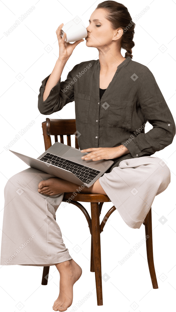Front view of a young woman wearing home clothes sitting on a chair with a laptop & drinking coffee