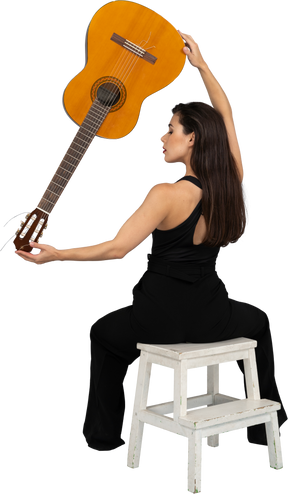 Back view of a young lady in black suit holding the guitar upside down and sitting on stool