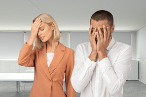 Блондинка в персиковой куртке с рукой на лбу и афро мужчина с руками, закрывающими лицо, стоят вместе в конференц-зале