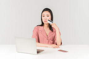Giovane donna asiatica pensierosa che tiene la carta di credito durante lo shopping online