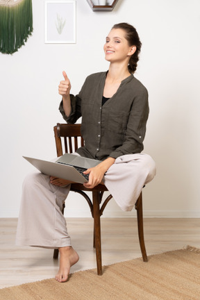 ノートパソコンで椅子に座って親指を上げて笑顔の若い女性の正面図