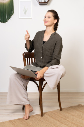 Front view of a smiling young woman sitting on a chair with a laptop & showing thumb up