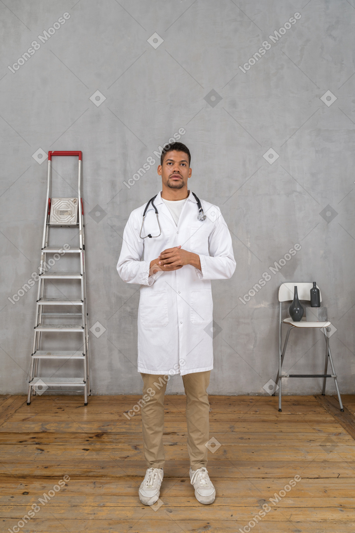 Front view of a young doctor standing in a room with ladder and chair holding hands together