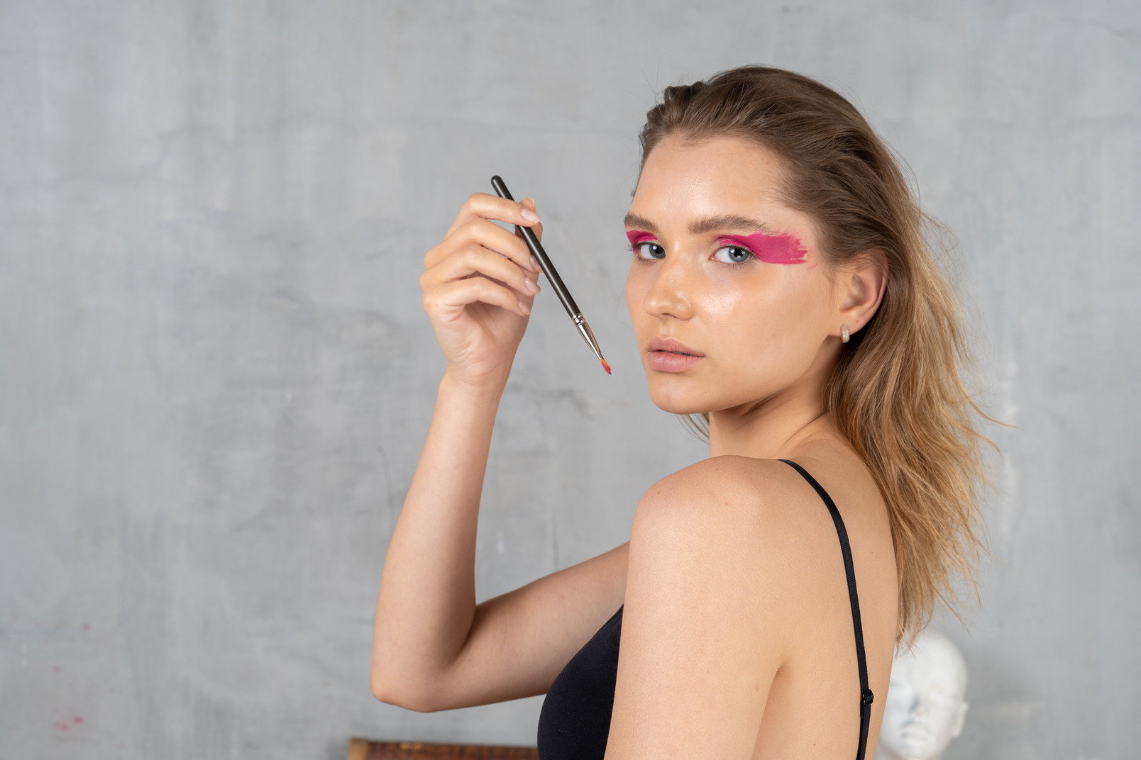 Side view of a young woman with bright pink eye make-up looking at camera while holding brush
