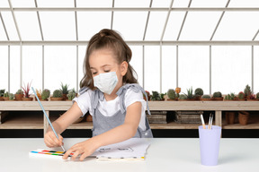 A little girl with mask sitting at a table and drawing