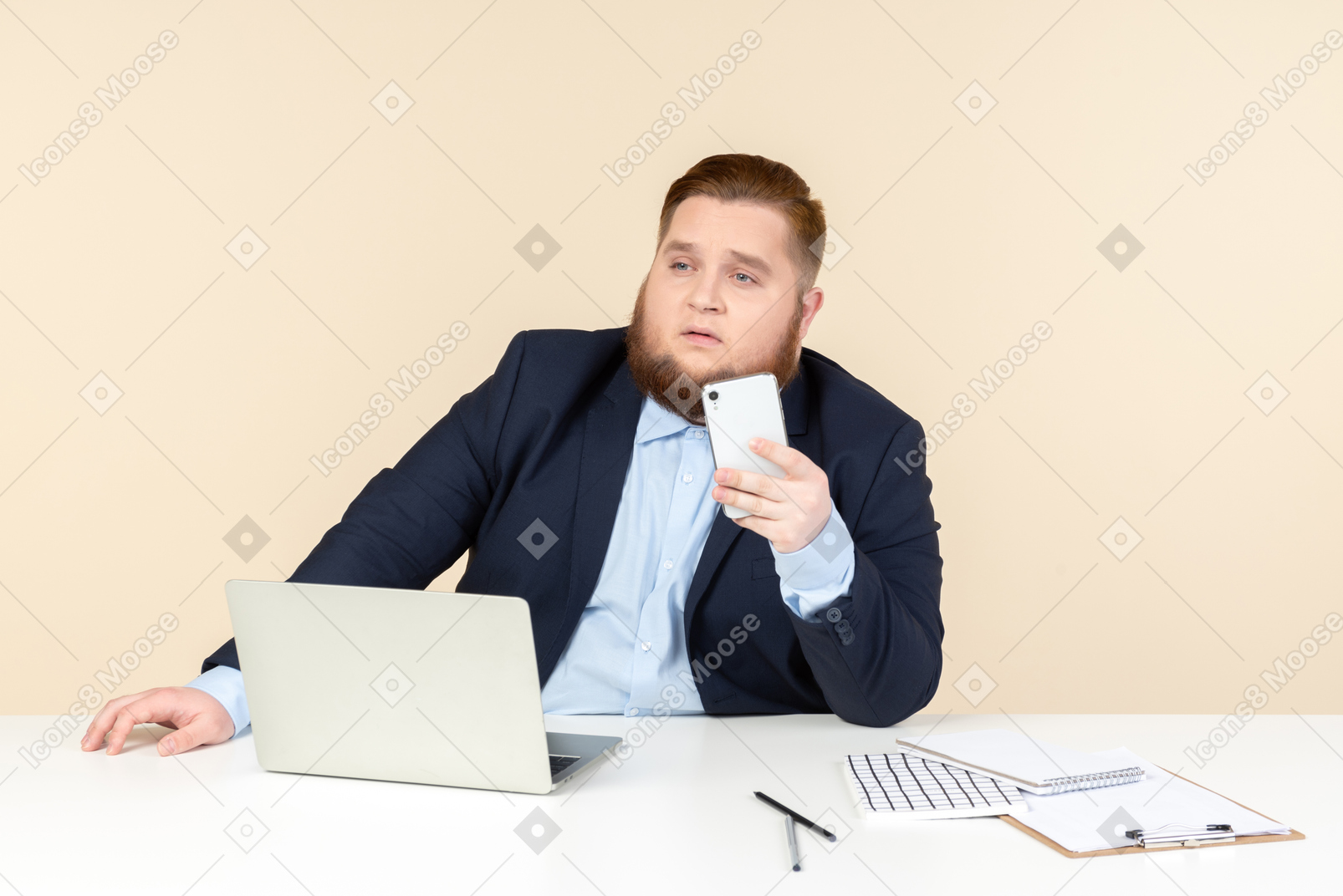 Задумчивый молодой человек с избыточным весом сидит за офисным столом и держит телефон