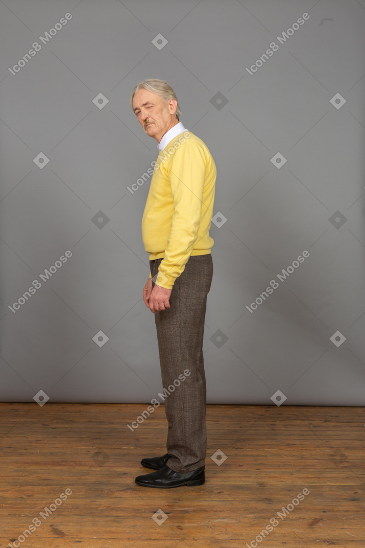 Vista lateral de um velho curioso em um pulôver amarelo virando a cabeça e piscando