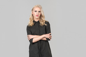 A young blond-haired person in a black dress  standing against the plain grey background