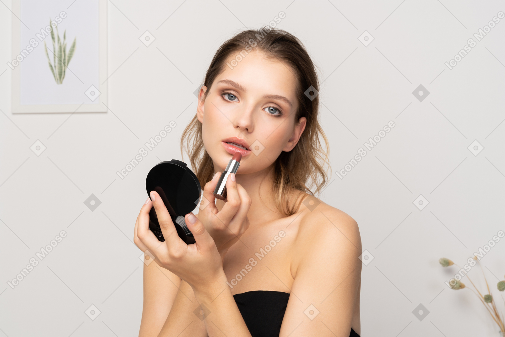 Front view of a young woman applying lipstick while holding a mirror