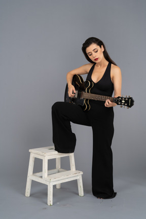 Vue de trois quarts d'une jeune femme en costume noir tenant la guitare et mettant la jambe sur le tabouret