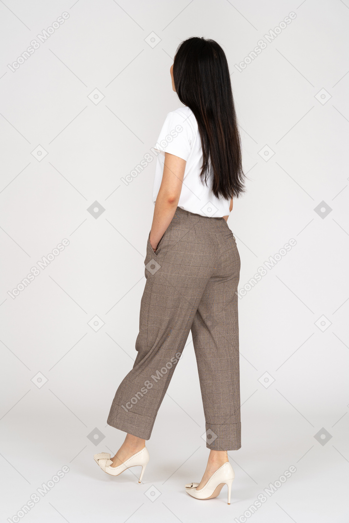 Vista posteriore di tre quarti di una giovane donna che cammina in calzoni e t-shirt mettendo la mano in tasca