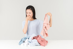 Young asian woman closing nose while holding some item from the pile on the table