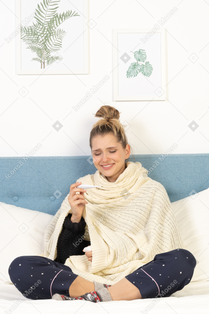 Front view of a smiling young lady wrapped in white blanket sitting in bed with thermometer
