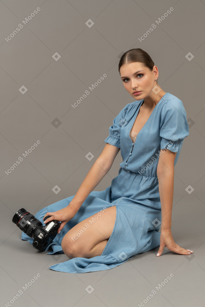 一位身穿蓝色连衣裙的年轻女子坐在地板上，拿着相机的侧视图