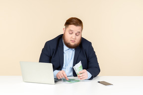 Молодой офисный работник с избыточным весом сидит за столом и считает деньги