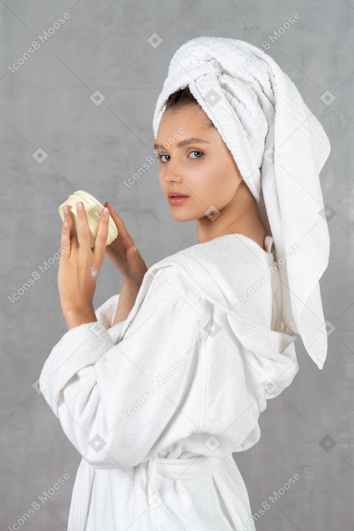 Vista lateral de uma mulher de roupão segurando uma banheira de creme
