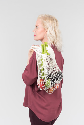 A nice-looking middle-aged blonde woman in a burgundy shirt holding close a shopper with some freshly bought vegetables
