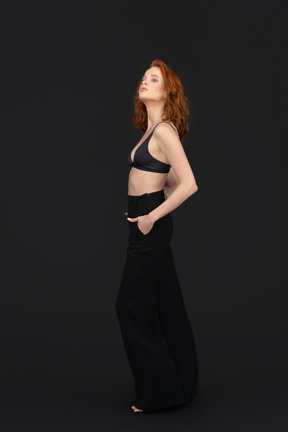 A side view of the sexy young woman standing on the black background holding her hands in the pockets