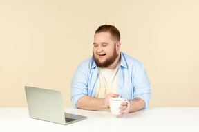 Смеющийся молодой мужчина с избыточным весом смотрит на ноутбук и пьет чай
