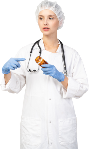 Vista frontal de una joven doctora señalando con el dedo el frasco de pastillas