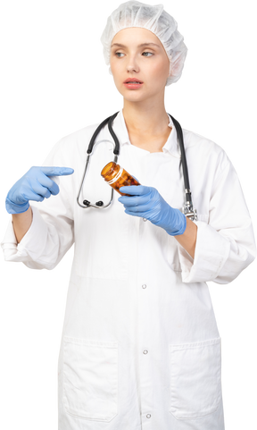 Front view of a young female doctor pointing finger at the jar of pills