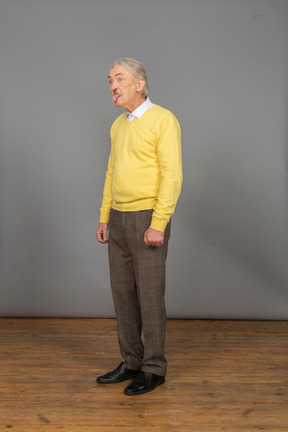 Vista di tre quarti di un vecchio curioso in pullover giallo che mostra la lingua e guarda da parte