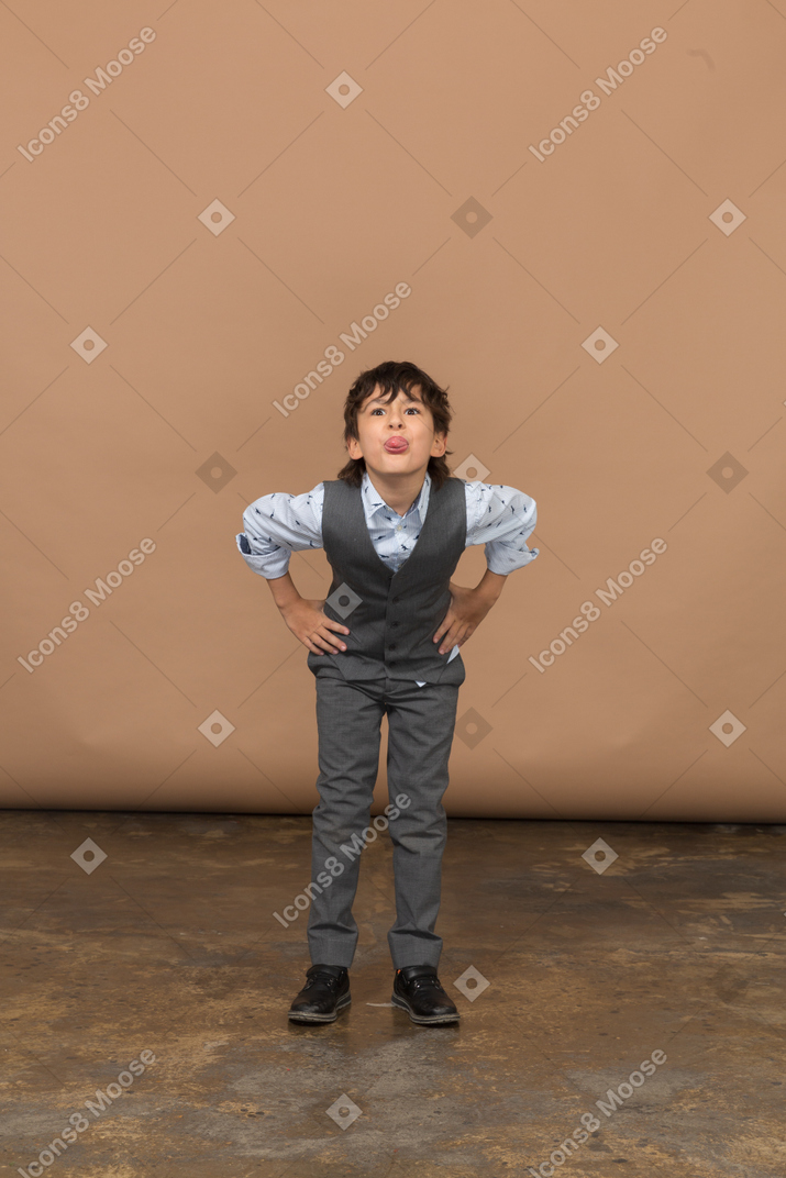 Вид спереди мальчика в костюме, стоящего с руками на бедрах и наклоняющегося вниз