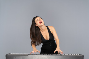 Front view of a pleased young lady in black dress playing the piano while singing