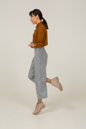 Vista laterale posteriore di una giovane donna asiatica che salta in calzoni e camicetta allargando le mani